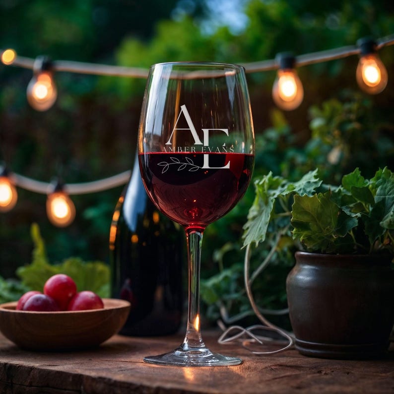 May include: A wine glass filled with red wine, featuring the text "AE AMBER EVANS" in white. The glass is on a wooden surface, with a bowl of grapes and a wine bottle in the background. Decorative lights and greenery add to the outdoor setting.