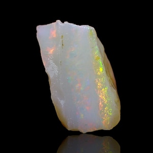 May include: A rough-cut opal gemstone with a milky white base color. The stone displays flashes of iridescent color, including red, orange, and green, across its surface. The opal is set against a black background.
