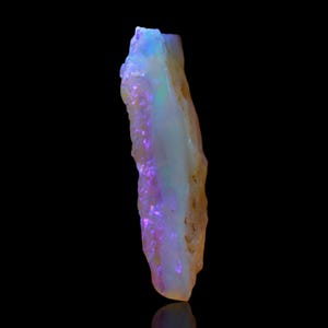 May include: A raw opal gemstone with a rough, irregular shape. The stone displays a mix of colours, including white, purple, blue, and yellow. The opal is set against a black background, highlighting its natural beauty.