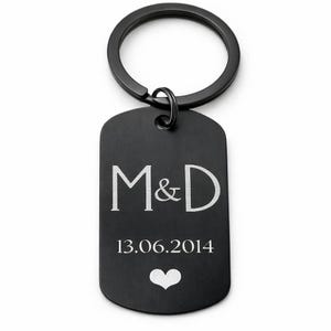 Op de afbeelding: Zwarte metalen sleutelhanger met een dog tag-vorm. De tag toont de initialen "M&D", de datum "13.06.2014" en een hartsymbool, allemaal in wit. De sleutelhanger heeft een zwarte ring.
