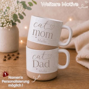 Könnte beinhalten: Zwei gestapelte Keramikbecher mit gesprenkeltem weißem Oberteil und beigefarbenem Boden. Der obere Becher trägt den Schriftzug cat mom und Mailo mit einem Herzsymbol. Der untere Becher trägt den Schriftzug cat Dad und Mailo mit einem Herzsymbol.