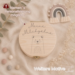 Könnte beinhalten: Eine runde Holzschatulle mit Klappdeckel, laser-graviertem Bärenmotiv und dem Schriftzug Meine Milchzähne. Die Box verfügt über einen Metallverschluss und ist mit einem kleinen dekorativen Regenbogen aus Garn sowie Filzkugeln arrangiert.
