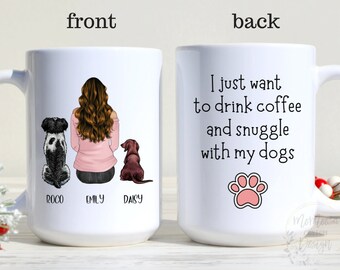 May include: White ceramic coffee mug with a personalized design featuring a woman sitting with two dogs. The text on the mug reads "I just want to drink coffee and snuggle with my dogs" and includes a pink paw print. The front of the mug has the names "Rocco", "Emily", and "Daisy" written below the illustration.