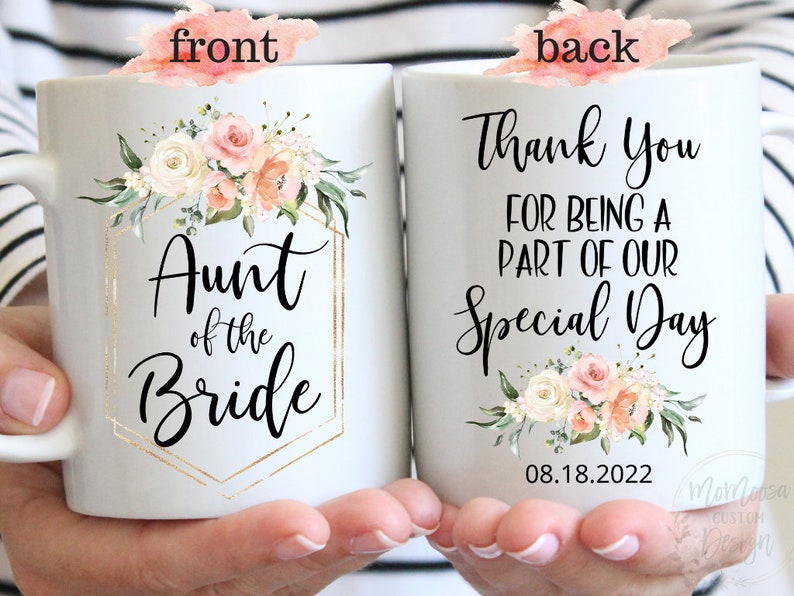 K&ouml;nnte beinhalten: Eine wei&szlig;e Keramiktasse mit einem Blumendesign und goldenen Akzenten. Die Vorderseite der Tasse zeigt den Text "Aunt of the Bride". Die R&uuml;ckseite der Tasse zeigt den Text "Thank You for being a part of our Special Day" und das Datum "08.18.2022".