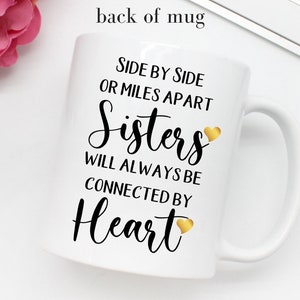 May include: White ceramic mug with black text that reads "Side by side or miles apart Sisters will always be connected by Heart" with a gold heart graphic after "Sisters" and "Heart".