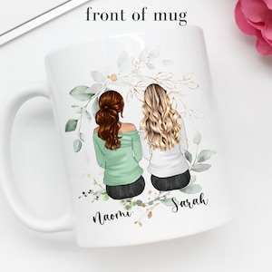 May include: White ceramic mug with a green and gold floral design. Two women with brown and blonde hair are depicted sitting back to back with the names "Naomi" and "Sarah" written below them.