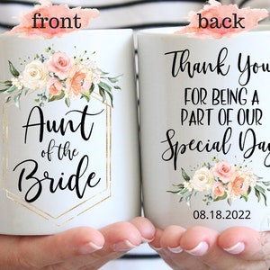 K&ouml;nnte beinhalten: Eine wei&szlig;e Keramiktasse mit einem Blumendesign und goldenen Akzenten. Die Vorderseite der Tasse zeigt den Text "Aunt of the Bride". Die R&uuml;ckseite der Tasse zeigt den Text "Thank You for being a part of our Special Day" und das Datum "08.18.2022".