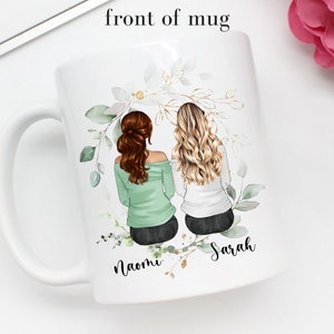 May include: White ceramic mug with a green and gold floral design. Two women with brown and blonde hair are depicted sitting back to back with the names "Naomi" and "Sarah" written below them.
