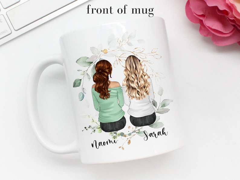 May include: White ceramic mug with a green and gold floral design. Two women with brown and blonde hair are depicted sitting back to back with the names "Naomi" and "Sarah" written below them.