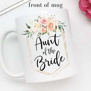 K&ouml;nnte beinhalten: Wei&szlig;e Keramiktasse mit goldenem geometrischem Rahmen und Blumenmuster. Der Text "Aunt of the Bride" ist in schwarzer Kursivschrift geschrieben.