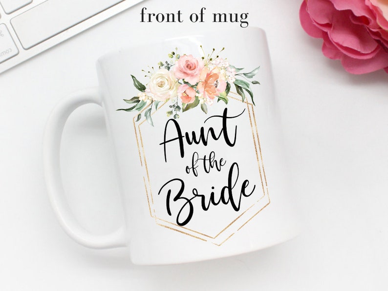 K&ouml;nnte beinhalten: Wei&szlig;e Keramiktasse mit goldenem geometrischem Rahmen und Blumenmuster. Der Text "Aunt of the Bride" ist in schwarzer Kursivschrift geschrieben.