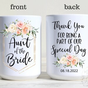 K&ouml;nnte beinhalten: Wei&szlig;e Keramiktasse mit einem goldenen geometrischen Rahmen und einem Blumendesign. Der Text "Aunt of the Bride" befindet sich im Rahmen. Die R&uuml;ckseite der Tasse hat den Text "Thank You for being a part of our Special Day" mit einem Blumendesign und dem Datum "08.18.2022".