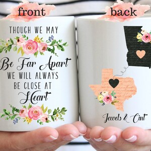 May include: White ceramic mug with a floral design and the text "Though we may be far apart we will always be close at heart". The back of the mug has a peach-colored outline of Texas with a black heart and a black outline of Georgia with a peach-colored heart. The states are connected by a dotted line. The text "Jewels & Cint" is printed below the states.