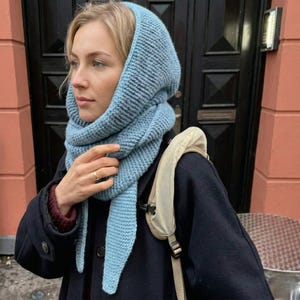 May include: A light blue knitted hood and scarf set. The hood is pulled up, framing the face, and the scarf is wrapped around the neck. The set is made of a chunky knit material. The person is wearing a dark coat.