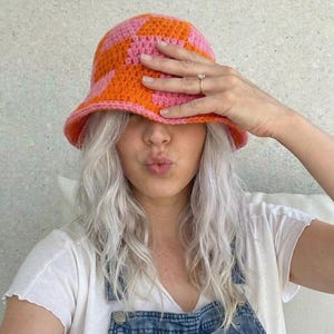 May include: A handmade crochet bucket hat in a checkerboard pattern of orange and pink. The hat is worn by a person wearing a white t-shirt and denim overalls. The hat's brim is a solid orange color.