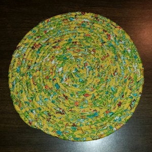 May include: A round yellow and green fabric trivet with a floral pattern. The trivet is made of braided fabric and has a circular shape.