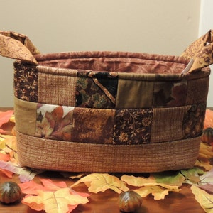 May include: Oval-shaped fabric basket with two handles, featuring a patchwork design in shades of brown, tan, and olive green. The interior lining is a patterned brown fabric. The basket is surrounded by fall leaves and small gourds.