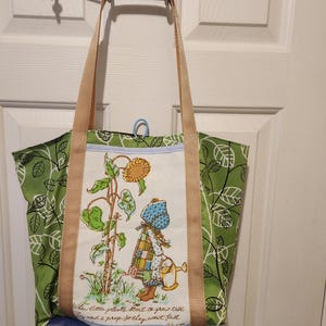 May include: Green tote bag with a floral pattern and beige straps. The central panel features a vintage illustration of a girl watering a sunflower. The bag has a blue base and a quote at the bottom.
