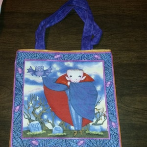May include: A Halloween-themed tote bag with a purple handle and trim. The bag features a cartoon vampire with a red cape and a bat against a starry night sky. The border has spiderwebs and bats. The bag is square-shaped.