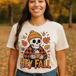 May include: A woman wearing a cream-colored t-shirt with a graphic of a skeleton in a beanie and plaid shirt holding a pumpkin spice latte. The shirt has the text "HEY FALL" in orange. She is also wearing a brown beanie and blue jeans.
