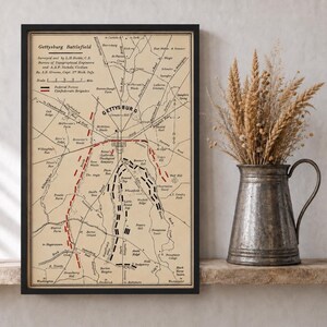 May include: A framed historical map titled Gettysburg Battlefield with black lines on a tan background, featuring red and black dashed lines indicating troop positions, a scale, and text detailing the survey by L.H. Biehle and A.A.P. Nichols.