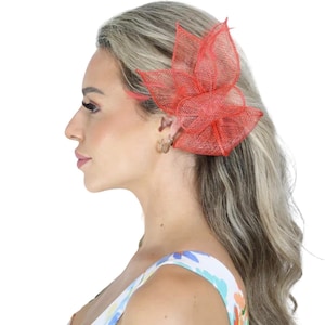 Puede incluir: Un accesorio para el cabello rojo en forma de flor, hecho de un material similar a una malla. El accesorio se usa en el costado de la cabeza. La persona lleva una blusa con estampado floral y aros dorados. El fondo es blanco.