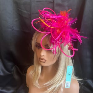 May include: A bright pink and orange feather fascinator, displayed on a mannequin head with blonde hair. The headpiece features decorative elements and is set against a dark backdrop. Ideal for events and special occasions.