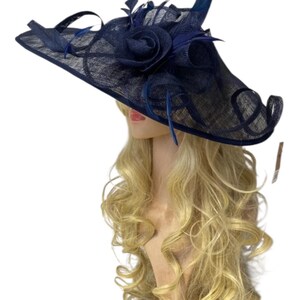 May include: A navy blue hat with a wide brim and decorative accents. The hat features a large, fabric rose and delicate feathers. The brim has a unique, looped design. The hat is displayed on a mannequin head with long, blonde hair.