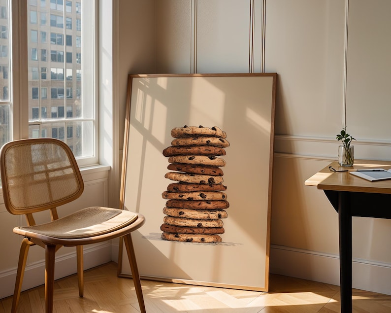 May include: A framed wall art print featuring a vertical stack of ten round chocolate chip cookies. The cookies are golden brown with visible dark chocolate chips, rendered in a detailed illustration style against a solid light beige background.