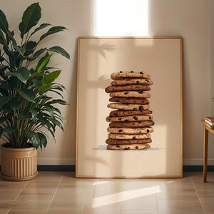 May include: A rectangular framed wall art print featuring a vertical stack of eleven golden-brown chocolate chip cookies centered on a solid light beige background. The cookies are round with visible dark chocolate chips and textured surfaces.