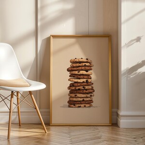 May include: A rectangular wall art print in a gold frame featuring a vertical stack of ten round chocolate chip cookies against a solid beige background. The cookies vary in shades of golden brown and dark brown, showing visible chocolate chips.