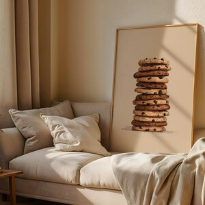 May include: A vertical rectangular framed wall art print featuring a centered illustration of a tall, precarious stack of eleven round chocolate chip cookies against a solid, light beige background, with visible dark brown chocolate chips.
