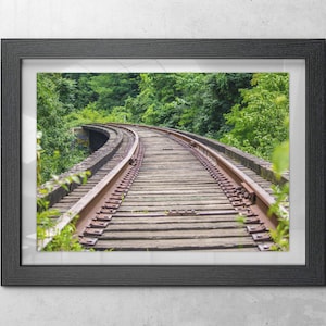 May include: Framed photograph of a curved railway track made of wooden planks and metal rails. Lush green trees surround the track, creating a natural tunnel effect. The image is framed in a black wooden frame.