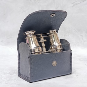 May include: A pair of polished brass opera glasses with a central focus wheel, housed inside a black leather case with a snap button closure. The binoculars feature two cylindrical barrels and a bridge mechanism, partially visible within the open case.