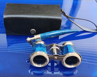 Könnte beinhalten: Vintage Operngläser mit blau-silbernem Finish, auf einer blauen Oberfläche. Die Gläser werden von einem passenden blauen Griff und einem schwarzen Lederetui mit Riemen begleitet.