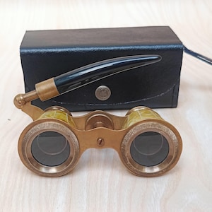 May include: Vintage opera glasses in brass with a black handle and a black leather case. The glasses feature a gold-coloured frame with detailed patterns around the lenses. The case is rectangular with a button clasp and a thin strap.