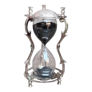 May include: An hourglass with a silver-colored metal frame and clear glass. The top chamber is filled with black sand, and the bottom chamber is empty. The hourglass has a decorative design and is likely used for timing.