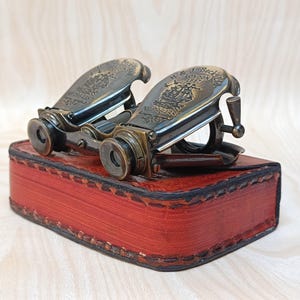 May include: Antique brass binoculars with the text "J. Beck London 1857" engraved on the top, resting on a red leather case. The binoculars have a detailed design and are a vintage item. The case is rectangular and stitched with dark thread.
