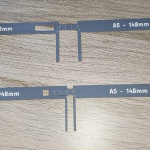 Può includere: Due strumenti di misurazione grigi, piatti e a forma di L con segni bianchi. Ogni strumento presenta il testo A5 - 148mm stampato sulla barra orizzontale. Le sezioni verticali includono scale numeriche da 0 a 4 e le sezioni orizzontali mostrano segni di misurazione.