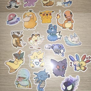 May include: A collection of colorful, glittery stickers featuring various Pokémon characters. The stickers include Pikachu, Charmander, Squirtle, and others, with the text "I Choose You" on one sticker. The stickers are on a light wood-grain surface.