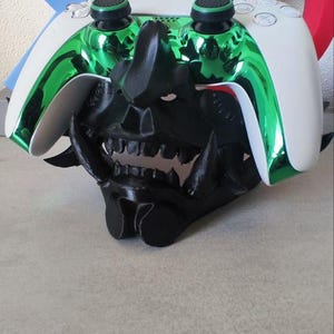 Peut inclure: Une manette PlayStation 5 personnalisée avec une finition chromée verte et des accents noirs, posée sur un masque Oni noir stylisé. La manette a des panneaux latéraux blancs et des joysticks noirs.