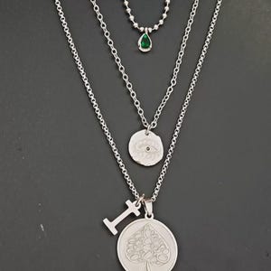May include: A layered silver necklace set. The top necklace features a small green teardrop pendant. The middle necklace has a circular pendant with an eye design. The bottom necklace has a spade pendant and a small letter "I" pendant.