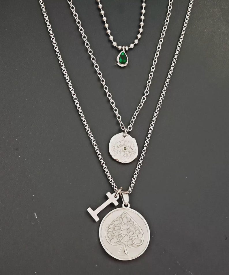 May include: A layered silver necklace set. The top necklace features a small green teardrop pendant. The middle necklace has a circular pendant with an eye design. The bottom necklace has a spade pendant and a small letter "I" pendant.