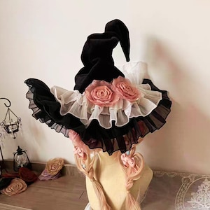 May include: A black witch hat with a velvet finish, featuring a wide brim with layers of black and white ruffles. Two large, pink fabric roses adorn the front. The hat is designed with a pointed crown.