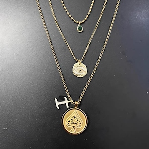 May include: A set of three gold-colored necklaces. The top necklace has a small green teardrop pendant. The middle necklace has a round pendant with an eye design. The bottom necklace has a spade pendant and a small letter "H" pendant.