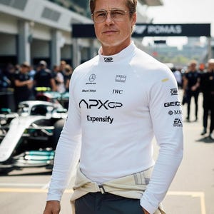 Könnte beinhalten: Ein weißes Langarm-Rennshirt mit mehreren Logos, darunter "APXGP" und "Expensify". Das Shirt wird mit einer dunklen Hose und einem hellfarbigen Rennanzug getragen. Die Person trägt eine Brille und steht vor einem Rennwagen.