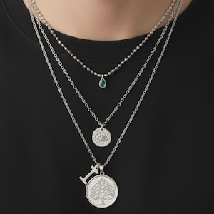 May include: A layered silver necklace set. The top layer features a small green teardrop pendant. The second layer has a small round pendant with an eye design. The bottom layer has a pendant with a spade and the letter "I".
