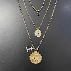 May include: Gold-toned layered necklaces. The top necklace has a small green teardrop pendant. The middle necklace has a round pendant with an eye design. The bottom necklace has a spade pendant and a small letter "H" charm.
