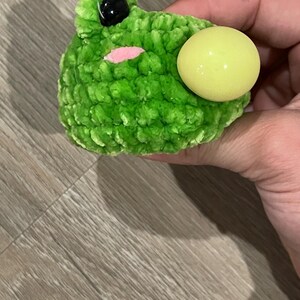 May include: A handmade green crochet frog with a yellow eye and black eye. The frog has a pink mouth and is made of a soft, textured yarn. The frog is a cute and whimsical toy.