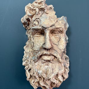 May include: A weathered, wall-mounted sculpture of a bearded man's face. The sculpture features a detailed beard and hair, with a color palette of tan, gold, and hints of blue. The artwork has a textured, aged appearance, suggesting an antique or classical theme.